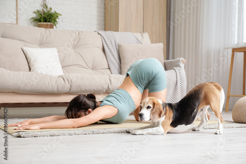 Young sporty woman with Beagle dog practicing yoga on mat in living room