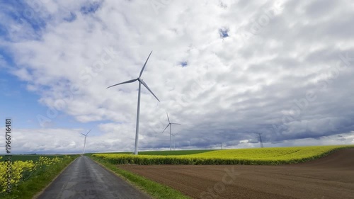 Rural landscape structured by contrasting tones where wind turbines rise above patchwork fields, with grey track cutting through yellow rapeseed and green crops beneath a layered cloudy sky