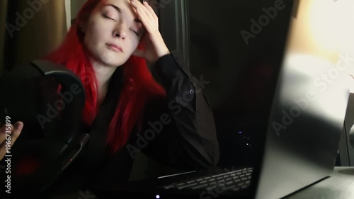 Caucasian woman organizes responsibilities during nighttime at her home workspace. She wears headset with microphone and starts to type on laptop looking at monitor. Lady looks tired reviewing tasks 