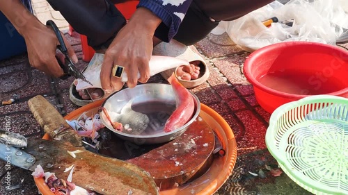 Hands of local vendor perform daily routine on street. Person uses scissors for fish scaling. Outdoor market features local vendor in daily routine. Scene captures authentic food process.