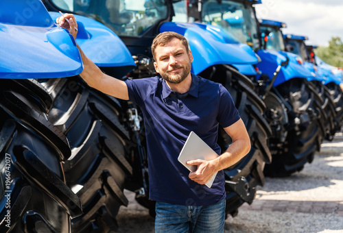 Modern male farmer holding tablet near tractors at farm equipment dealership. 