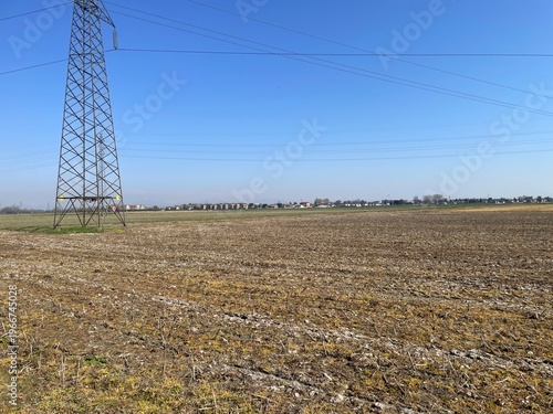 Power Line Over Horizon: A towering power line against a vast, open field, with a distant cityscape. Illustrating the interplay between technology, infrastructure, and the natural landscape.