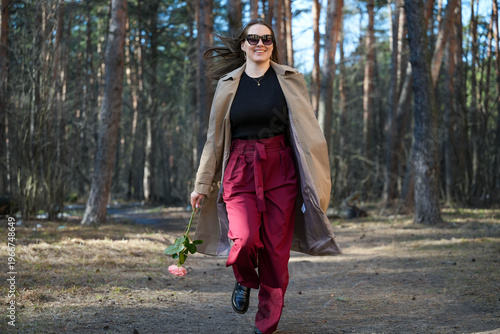 Tablou pe pânză Woman Caucasian Striding Through Woods With Bouquet In Hand, Trench Coat Open Re