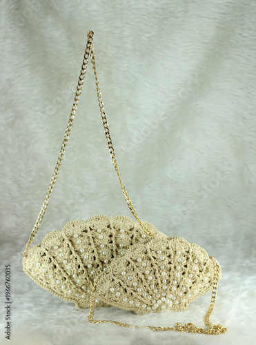 Beige crocheted shoulder bag in the shape of a seashell with a gold-colored handle sits on a white fluffy blanket. The bag features pearl bead details.