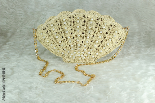 Beige crocheted shoulder bag in the shape of a seashell with a gold-colored handle sits on a white fluffy blanket. The bag features pearl bead details.	