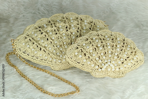 Beige crocheted shoulder bag in the shape of a seashell with a gold-colored handle sits on a white fluffy blanket. The bag features pearl bead details.	