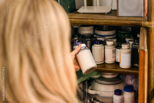 Blonde woman organizing bottles with food supplements and vitamins on kitchen shelf. Woman selecting supplement bottle from cabinet filled with vitamins and medicine containers at home