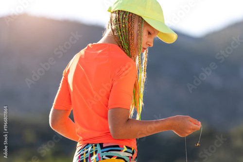 Young teenager girl colorful braids wear cap orange rashguard standing sea looking small crab fishing line. Summer seaside lifestyle scene teen child explore nature vacation Adriatic coast Croatia