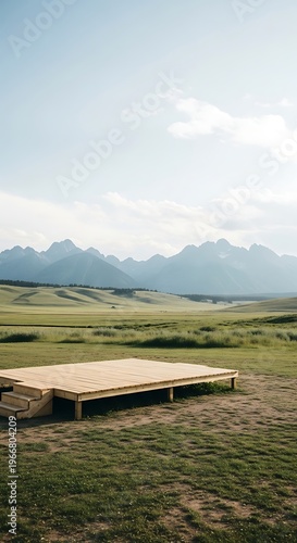 Wooden stage platform in a vast green meadow with majestic mountain range backdrop under a clear blue sky with soft clouds