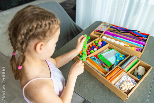 Little girl making craft with colorful materials at table using creative kit at home. Kids creativity development fine motor skills and learning through play concept.