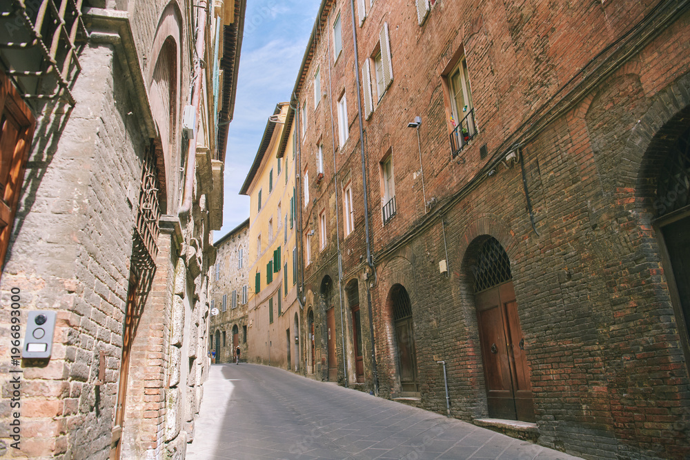 Fototapeta premium Narrow street in Siena Italy with old brick buildings and historic architecture, quiet European town scene under blue sky, travel destination with medieval charm and peaceful urban view