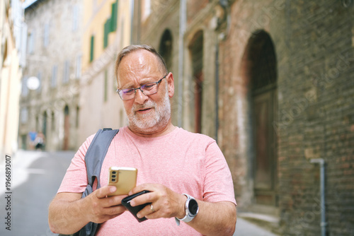 Senior man using smartphone while walking on quiet old street, wearing glasses and casual clothes, checking messages and holding wallet, modern lifestyle and daily activity concept