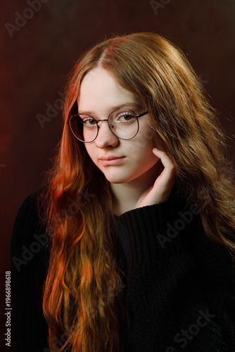 Moody studio portrait of a beautiful teenage girl with long ginger hair wearing round glasses and stylish black sweater on dark studio background.