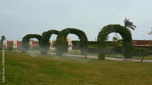 Mystic Garden Scene. Misty Topiary Arches Alongside Cityscape And Playful Sprinkler Effects