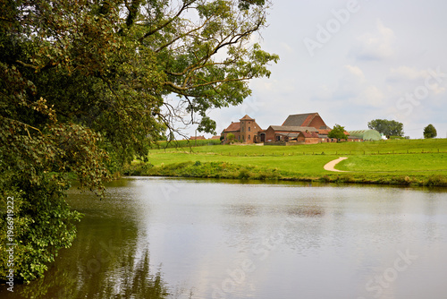 Serene rural landscape with a tranquil lake, lush green fields, charming farm buildings, and a winding path under a soft sky, evoking peaceful countryside charm.
