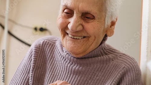 Senior woman with wrinkled skin and gray hair sitting at table smiling happily and sincerely surrounded by love and care. Her eyes shine with kindness and joy. Care and love for lonely elderly