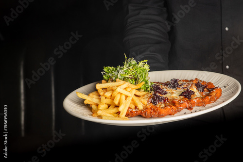 Fotografie A waiter holding a white plate of chicken parmigiana topped with melted cheese and beetroot, served with golden french fries and a side salad against a dark background