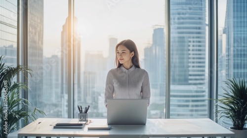 Asian businesswoman stands by window with laptop, looking at modern city skyline from her office.