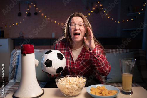 female football fan holding ball watching soccer game on tv at home sitting on sofa with beer and snacks screaming with hands gesture near mouth. Sport, leisure and entertainment
