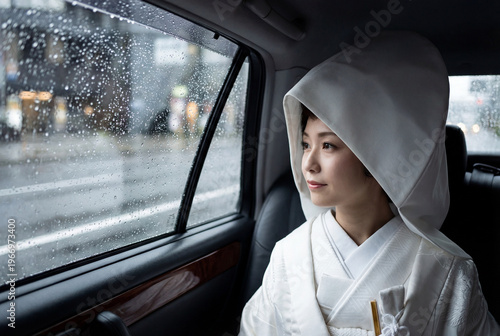 Japanese bride in a traditional white kimono and wataboshi hood sits inside a car, looking thoughtfully out the rain-streaked window.