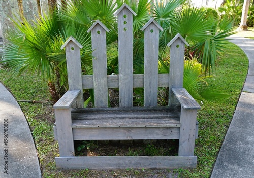 A whimsical, weathered wooden bench with a tall birdhouse-shaped backrest sits on grass beside a sidewalk and palm plants at Palma Sola Botanical Park, Bradenton, Florida, U.S