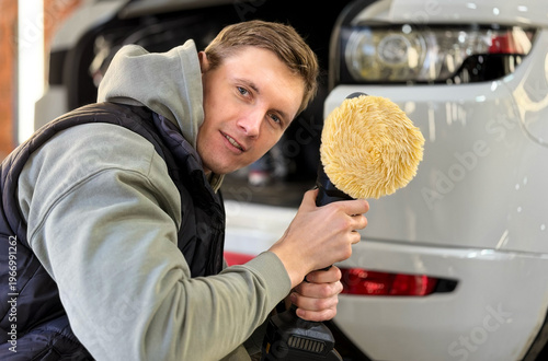 A man holding a car polishing machine with a fur circle in his hands looks at the camera and smiles against the blurred background. Detailing and polishing of the car. A service worker polishes car. 