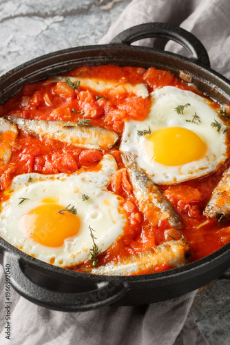 Fried eggs with canned sardines and vegetables simmered in tomato sauce close-up in a frying pan on the table. Vertical