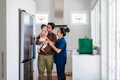 A kiwi father gently kissing their child with wife standing close to them in the kitchen