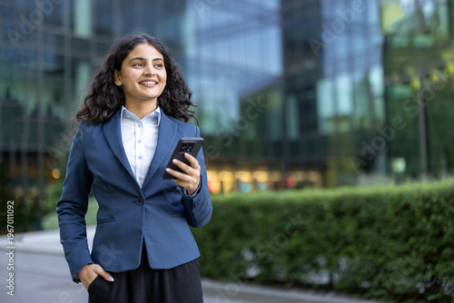 Young professional businesswoman smiling and holding a smartphone. Looking away in a modern urban environment with a glass office building in the background. Representing success and future endeavors