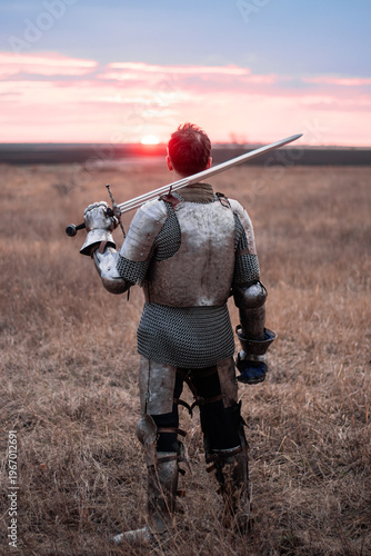 Canvastavla A knight in cuirass and chainmail, holding a sword and a bloodied face, stands with his back to the sunset in a field