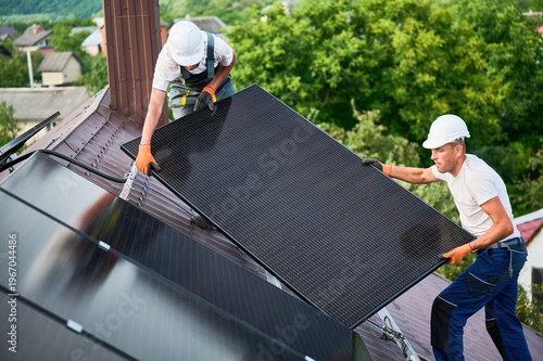 Workers building solar panel system on roof of house. Two men installers in helmets carrying photovoltaic solar module outdoors. Alternative, green and renewable energy generation concept.
