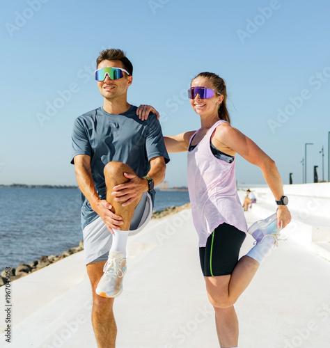 Athletic man and woman stretch legs by the waterfront promenade on a bright sunny day, preparing for exercise