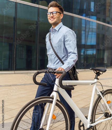 Smiling professional man in glasses walks a white bicycle outdoors near a modern glass office building during daytime