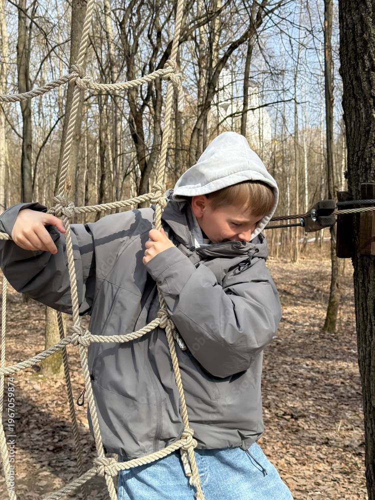 Naklejka premium Little Boy Playing in Rope Cargo Net Obstacle Course in Spring Forest Adventure Park Close Up
