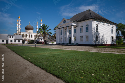 View of the Neveh Shalom Synagogue and the Ahmadiyya Anjuman Isha'at Islam Mosque standing side by side under a clear blue sky in Paramaribo, Paramaribo District, Suriname.