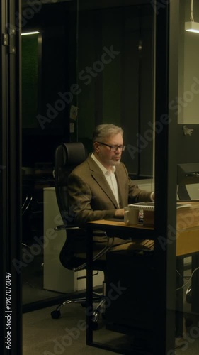 Wide vertical shot of stressed middle-aged Caucasian man working on report or project at desk in office at night, staring at computer screen, taking off glasses, massaging face and drinking coffee