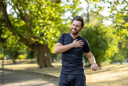 A young male athlete, twisted in pain, stands in the park and holds his hand on his shoulder