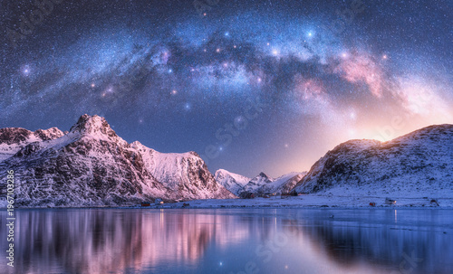 Milky Way arch above sea coast and snow covered mountains in winter at night. Lofoten Islands, Norway. Arctic landscape with blue starry sky, arched milky way reflected in water, snowy rocks. Space
