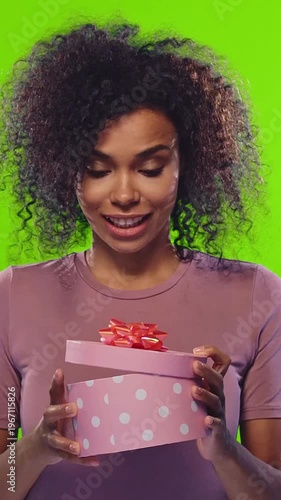 Happy young African American woman with curly hair opening round pink gift box with white polka dots and red ribbon on green background showing excitement and surprise. Celebration, present, happiness