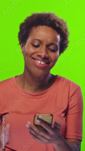 Young African American woman with curly hair wearing orange shirt using smartphone and smiling on green background. Technology, communication, mobile device and positive emotions concept.