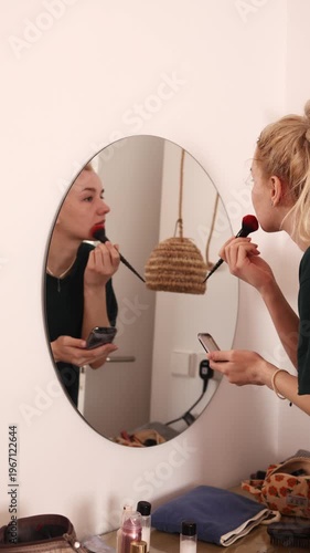 Young blonde woman applying lipstick with brush in front of round wall mirror at home interior. Perfect for beauty, cosmetics, makeup routine and lifestyle themed projects