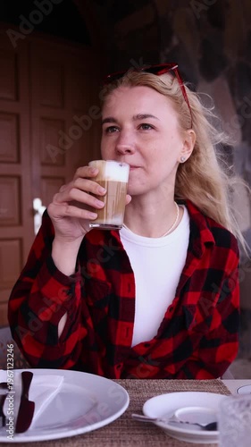 Young blonde woman drinking latte coffee at outdoor cafe table. Perfect for lifestyle, coffee, cafe culture, morning routine, travel and relaxation themed commercial projects