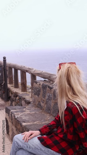 Back view of young blonde woman in red jacket gazing at misty Atlantic ocean from stone viewpoint with wooden railing in Tenerife. Perfect for travel and lifestyle projects