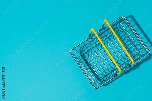 Flat lay of empty metal shopping basket with yellow handles on bright turquoise blue background. Concepts of shopping and sale season.