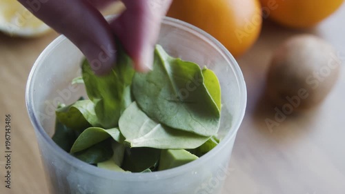 Close-up of a hand placing fresh fruit slices like kiwi and banana with green leaves into a glass, preparing a healthy infused water or smoothie. Natural detox drink being assembled on wooden kitchen.