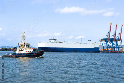 Car carrier ship docked at port terminal in Brazil being loaded with vehicles, while tugboat passes nearby, maritime logistics concept.