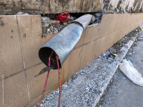 Metal ventilation pipe protruding from exterior building wall with exposed electrical wires and construction debris on ground