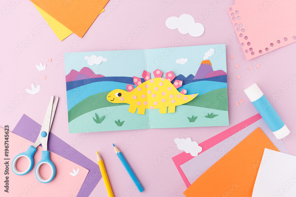 Obraz premium Paper cut dinosaur in landscape with mountains and volcano on folded card with scissors, glue stick, pencils and colored paper on pink background. Flat lay with copy space. Kids craft concept. 