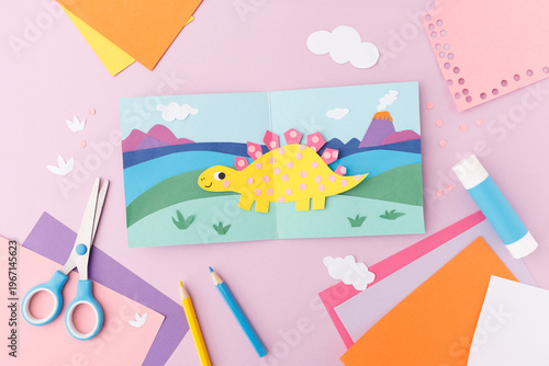 Paper cut dinosaur in landscape with mountains and volcano on folded card with scissors, glue stick, pencils and colored paper on pink background. Flat lay with copy space. Kids craft concept. 