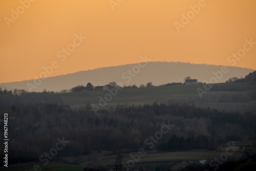Wieczór zapada nad Górami Świętokrzyskimi w ostatnich dniach zimy w Polsce, rzucając ciepły, pomarańczowy odcień. Krajobraz charakteryzuje się łagodnymi wzgórzami i drzewami, oraz domami